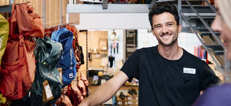 A smiling male salesperson in an outdoor store stands near colorful backpacks and helps a customer.