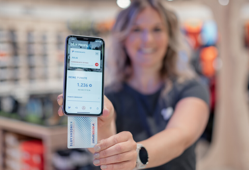 A woman in a retail store holds a smartphone, displaying the account balance of a digital wallet and a customer card.