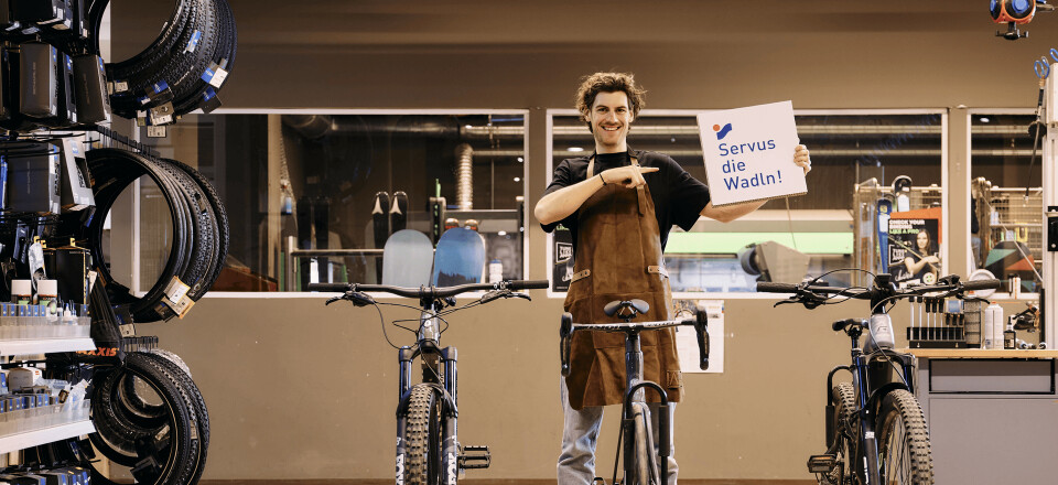 Ein lächelnder Fahrradmechaniker hält ein "Service op Maat!"-Schild und steht zwischen zwei Fahrrädern.