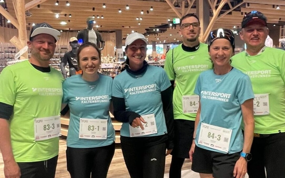 Sechs lächelnde Intersport-Teammitglieder in Sportshirts und Startnummern in einem Sportartikelgeschäft.