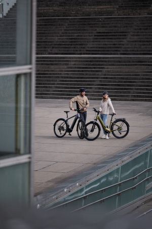 Paar mit zwei E-Bikes auf einem modernen Stadtplatz, Betonstufen und Glasarchitektur.