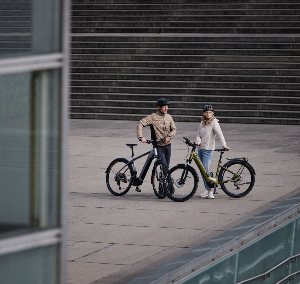 Couple with two e-bikes on a modern city square, concrete stairs and glass architecture.