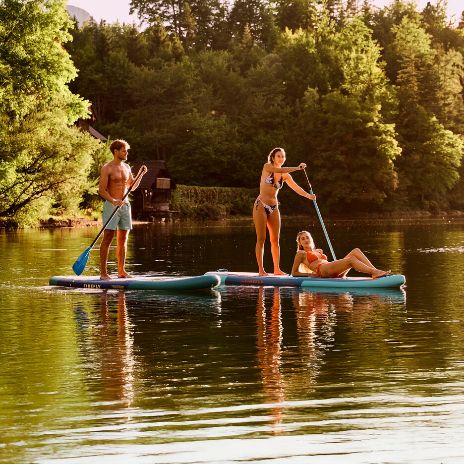 Drei Personen stehen auf Stand-Up-Paddleboards auf einem ruhigen See, der von einem dichten Wald umgeben ist, im warmen Abendlicht.