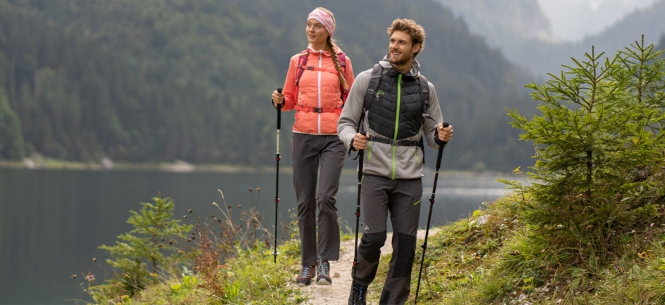 Ein lächelndes Paar wandert auf einem Wanderweg an einem Bergsee, gekleidet in Outdoor-Ausrüstung und mit Trekkingstöcken.