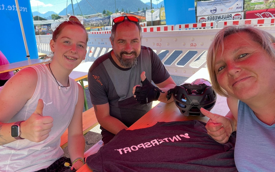 Drei lächelnde Personen, zwei Frauen und ein Mann, die bei einer Outdoor-Veranstaltung die Daumen hochhalten, mit einem Intersport T-Shirt und Helm.