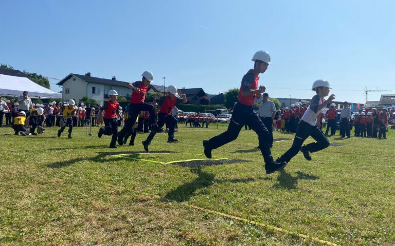 Jugendfeuerwehrleute in Helmen und Uniformen sprinten während eines Outdoor-Wettbewerbs über ein Grasfeld.