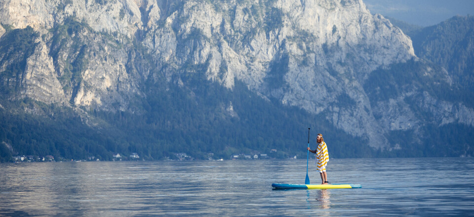 Eine Person beim Stand-up-Paddling auf einem ruhigen See mit majestätischen, baumbedeckten Bergen im Hintergrund.