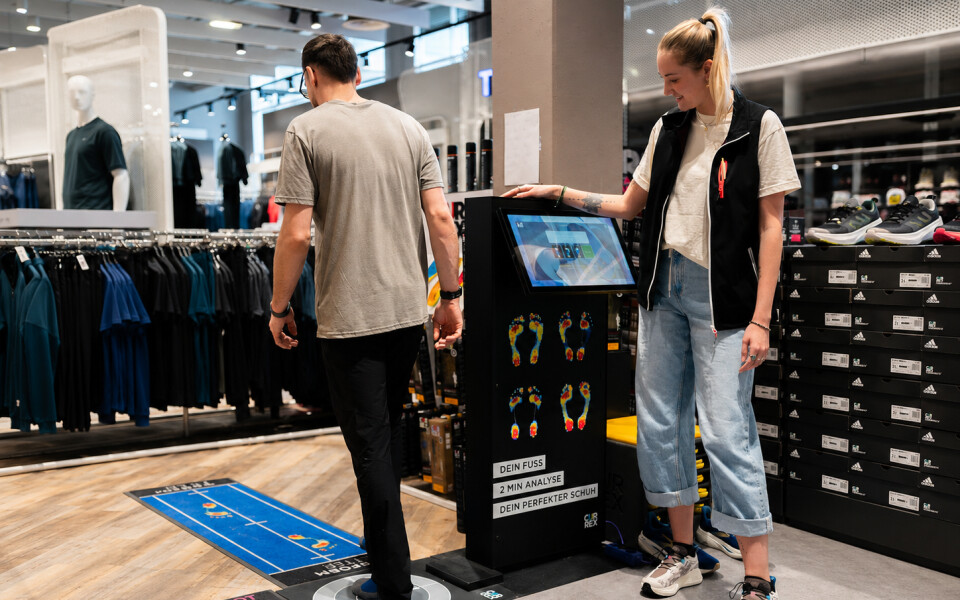 Der Kunde nutzt einen Currex Fußscanner für die personalisierte Schuhpassformbestimmung in einem Sportfachgeschäft, unterstützt von Mitarbeitern.