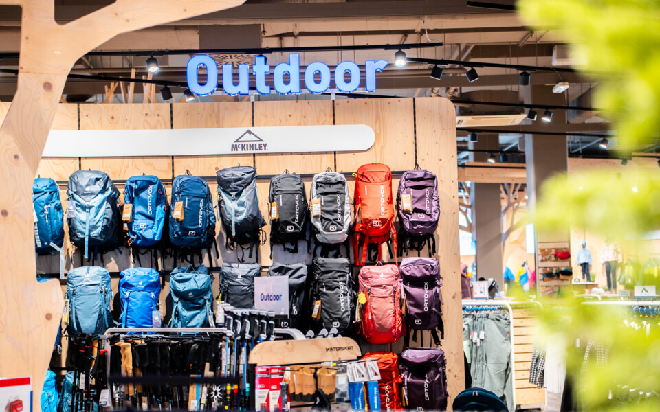 Schaufenster eines Outdoor-Ausrüstungsgeschäfts mit Wanderrucksäcken, Sportbekleidung und einem auffälligen "Outdoor"-Schild.