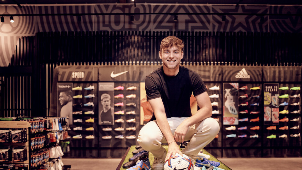Ein lächelnder Mann hockt neben einem Fußball und bunten Fußballschuhen in einem modernen Sportfachgeschäft.