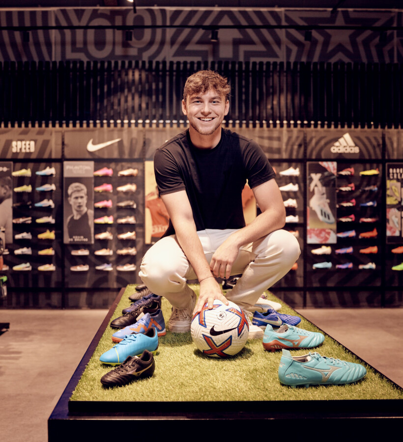 Lächelnder Mann hockt neben einem Fußball und bunten Fußballschuhen in einem modernen Sportfachgeschäft.