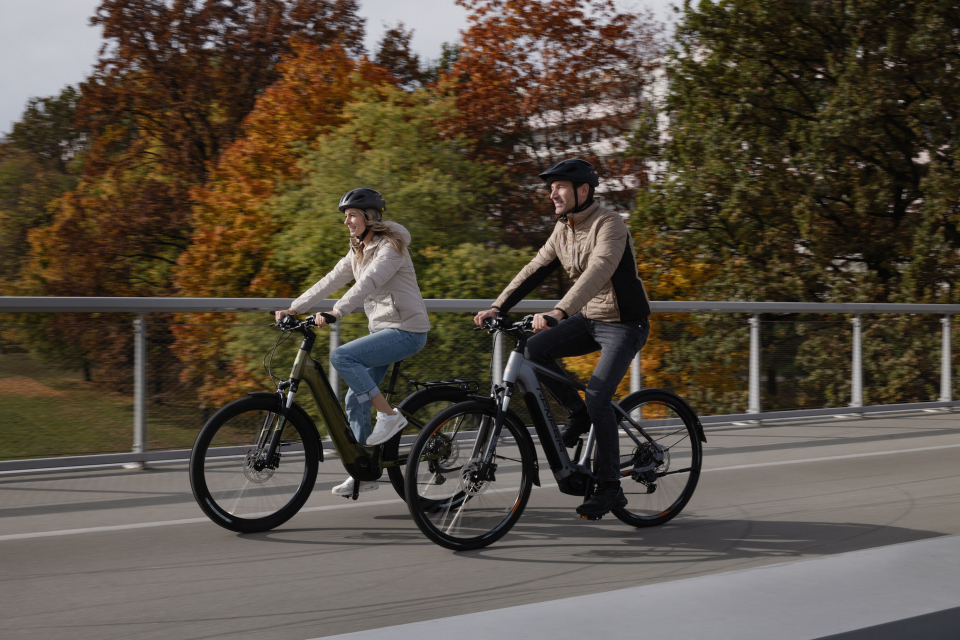 Mann und Frau fahren E-Bikes auf einer Brücke, umgeben von bunten Herbstbäumen.