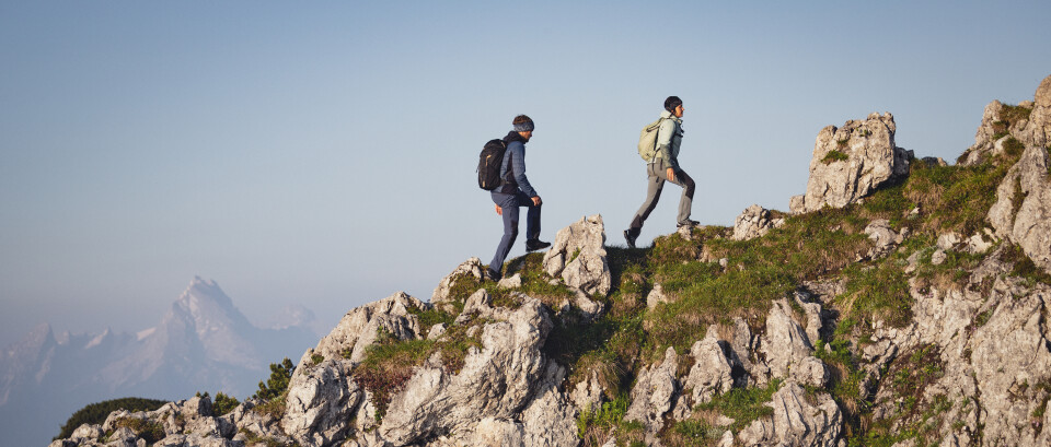 Zwei Wanderer mit Rucksäcken erklimmen einen schroffen Bergkamm und bewegen sich dabei durch felsiges Gelände und grüne Hänge mit fernen Gipfeln.