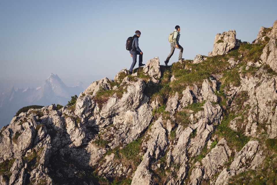 Two hikers with backpacks are climbing a rugged mountain ridge, moving through rocky terrain and green slopes with distant peaks.