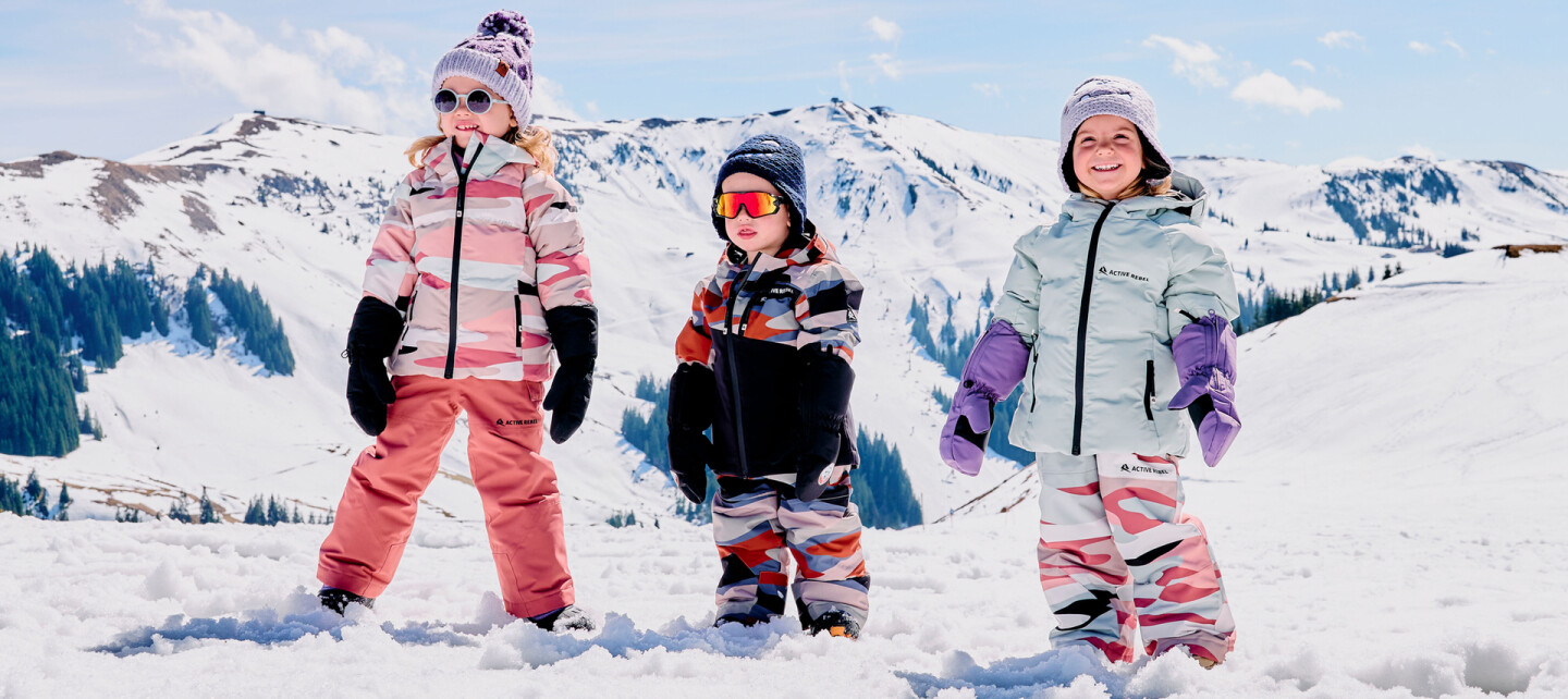 Drei Kinder in bunter Skibekleidung stehen im tiefen Schnee vor sonnigen Berggipfeln.