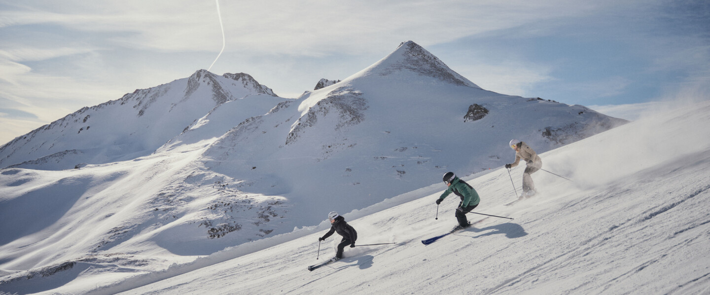 Drei Skifahrer beim Wintersport auf einem sonnigen, schneebedeckten alpinen Berghang.