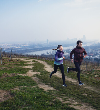 Ein Paar beim Trailrunning durch einen Weinberg mit Panoramablick auf die Stadt und einem Fluss darunter.