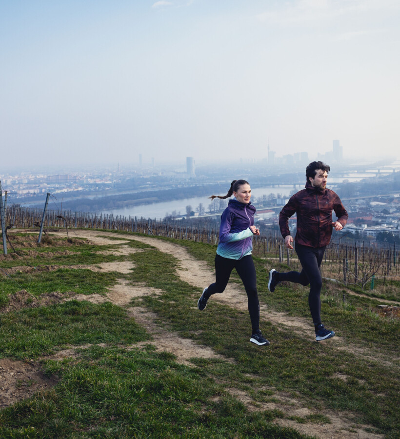 Ein Paar beim Trailrunning durch einen Weinberg mit Panoramablick auf die Stadt und einem Fluss darunter.